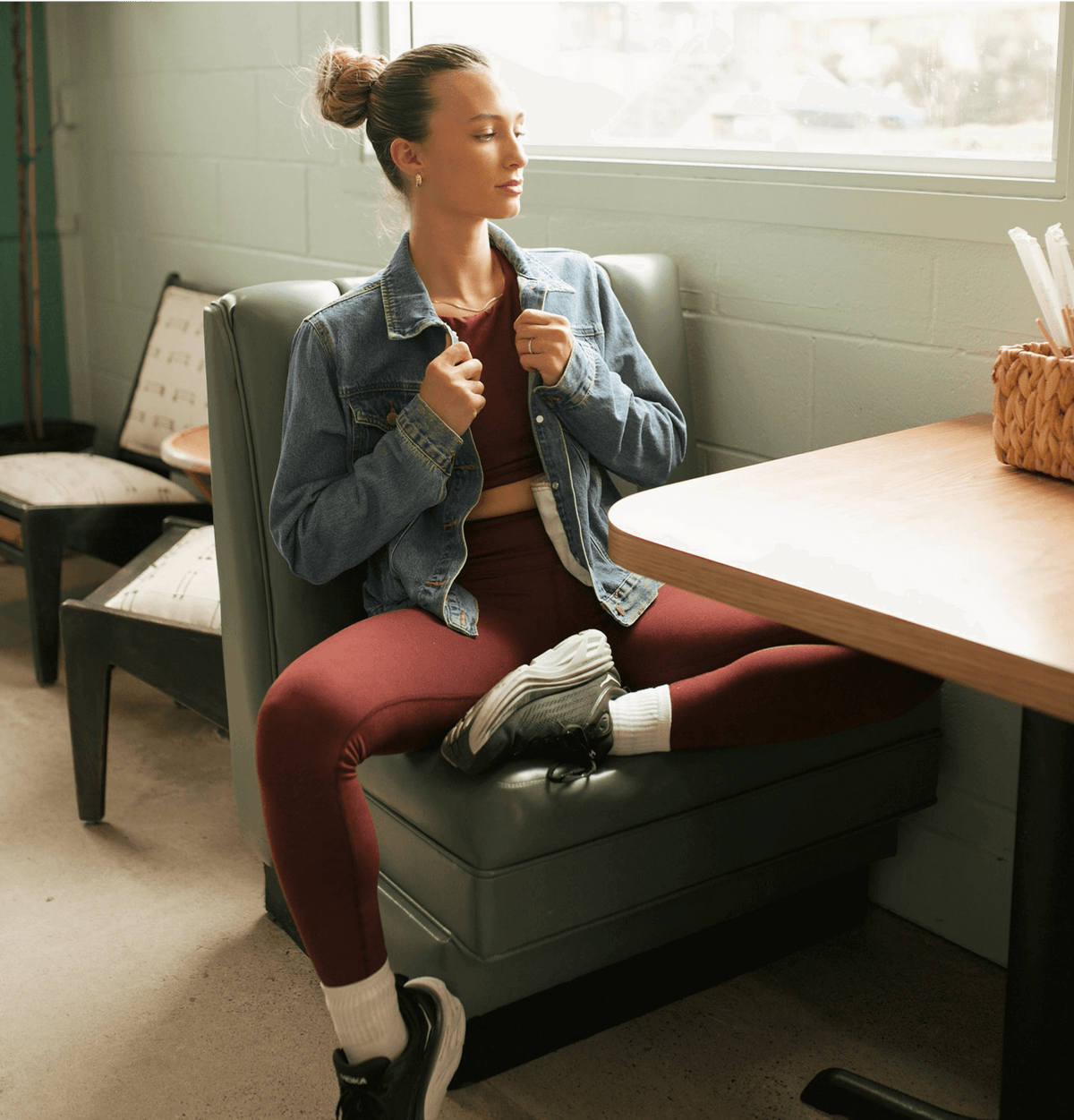 Woman sitting on a booth in a casual setting, wearing a denim jacket and burgundy leggings.