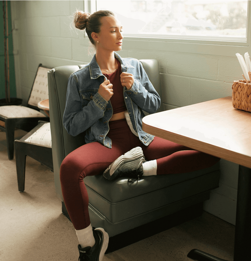 Woman sitting on a booth in a casual setting, wearing a denim jacket and burgundy leggings.